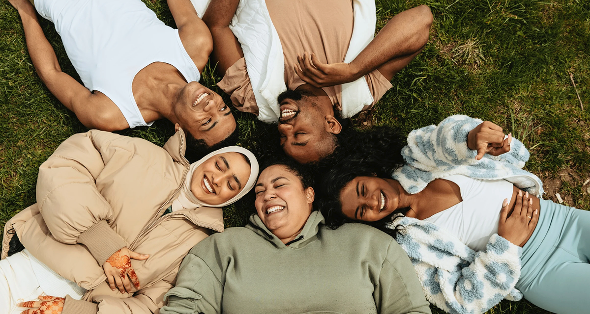 Diverse group of six friends lying in a circle on the grass, heads together, smiling and enjoying a sunny day.