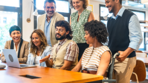 Diverse group of colleagues gathered around a laptop, smiling and collaborating at a bright coworking space.