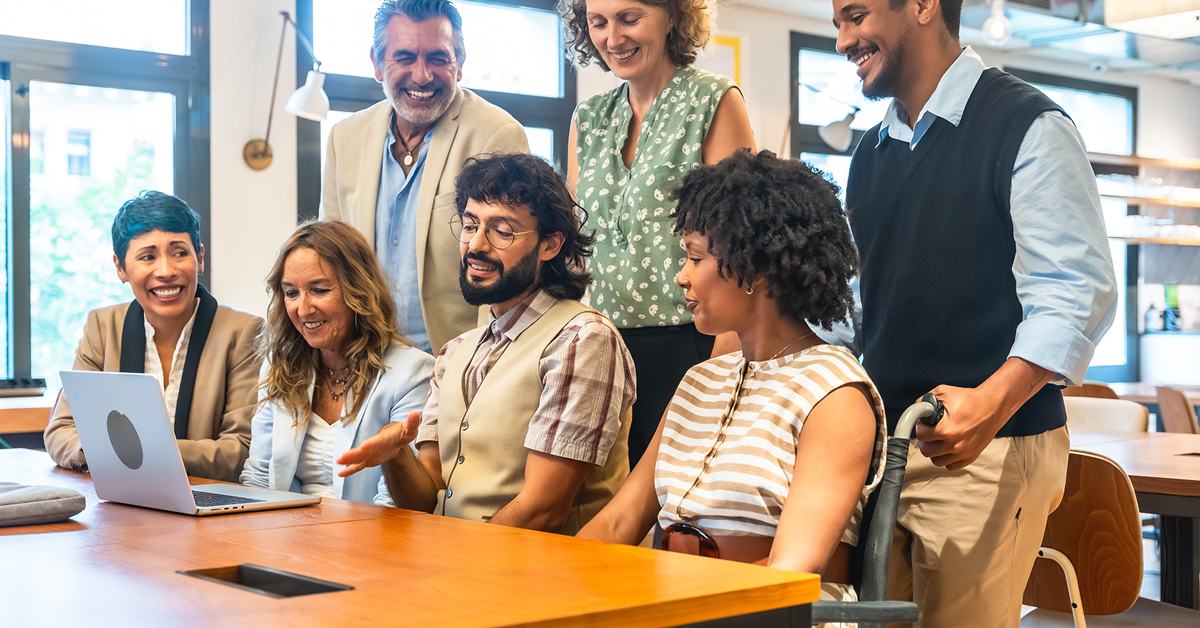 Diverse group of colleagues gathered around a laptop, smiling and collaborating at a bright coworking space.