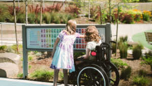 Two young girls at an outdoor park examine a large colorful information panel; one girl uses a wheelchair beside her friend.