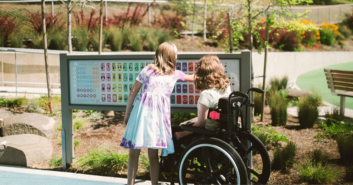 Two young girls at an outdoor park examine a large colorful information panel; one girl uses a wheelchair beside her friend.